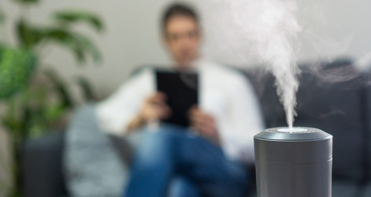 grey best buy humidifier with steam shown in focus in front of man reading on couch