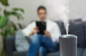 grey best buy humidifier with steam shown in focus in front of man reading on couch