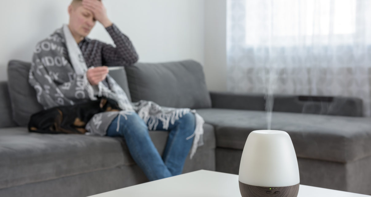 man with cold taking temperature on couch with dog next to humidifier